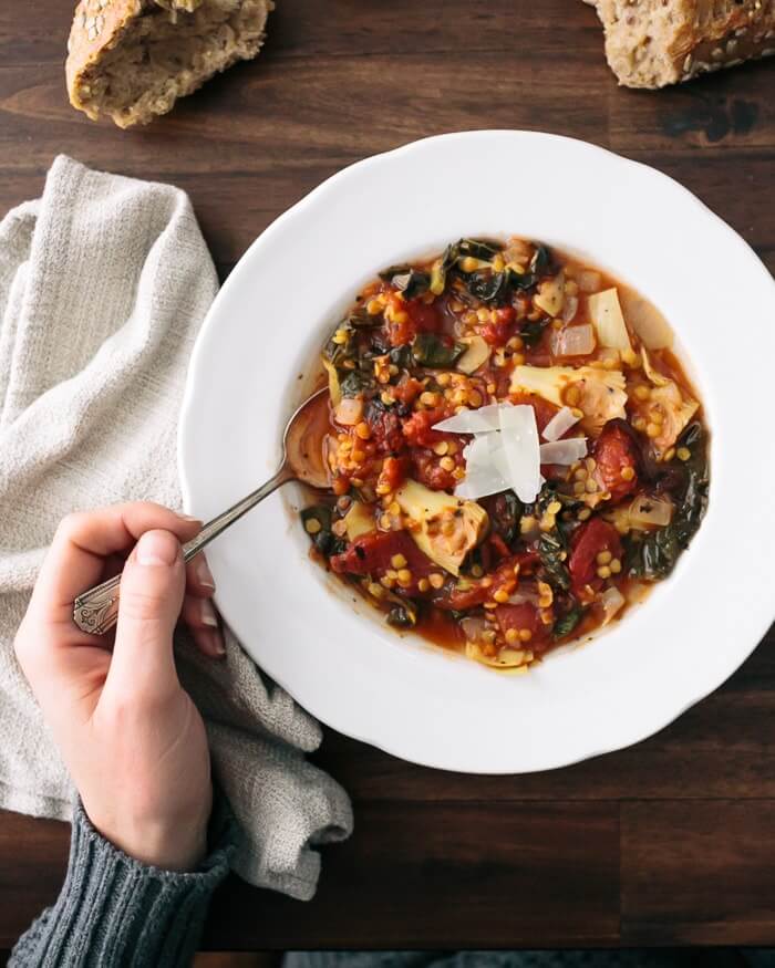 Italian lentil soup in a white bowl with   a hand holding a spoon in the bowl