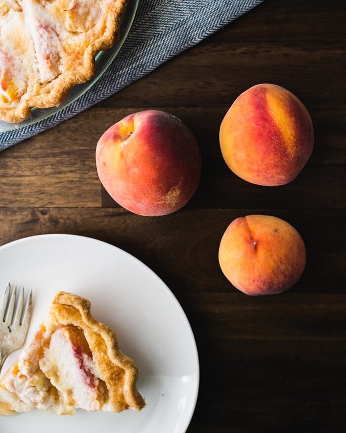 slice of homemade peach pie on white plate next to fresh peaches