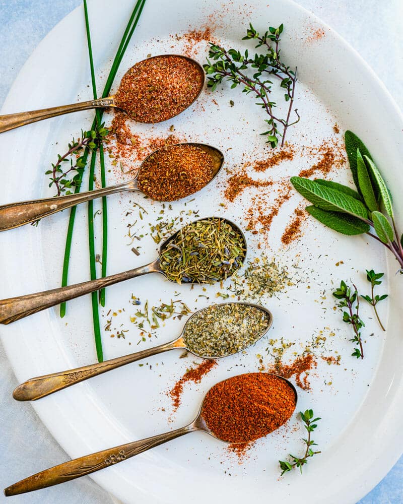 4 spoons lined up over a plate with different kinds of seasonings filling each spoon. An orange blend, and leafier blend, a grainy brown blend, and a red blend.