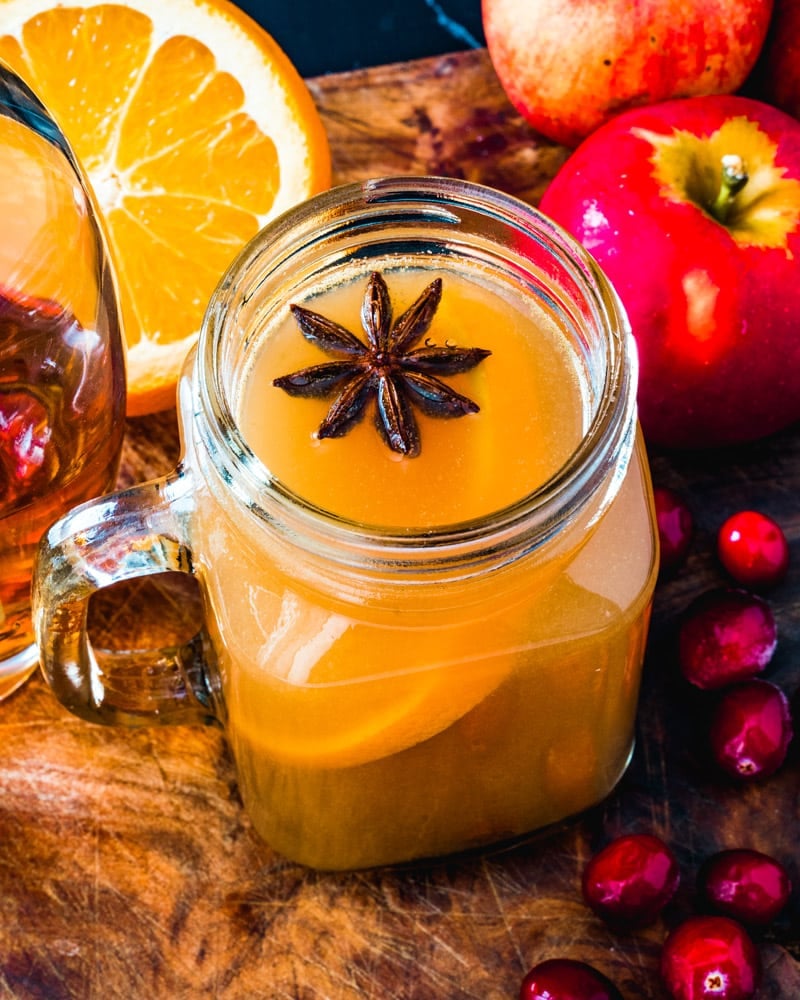 Spiked Apple Cider in a mason jar glass with a star anise floating in the drink, surrounded by fruits, and sitting on a wooden table