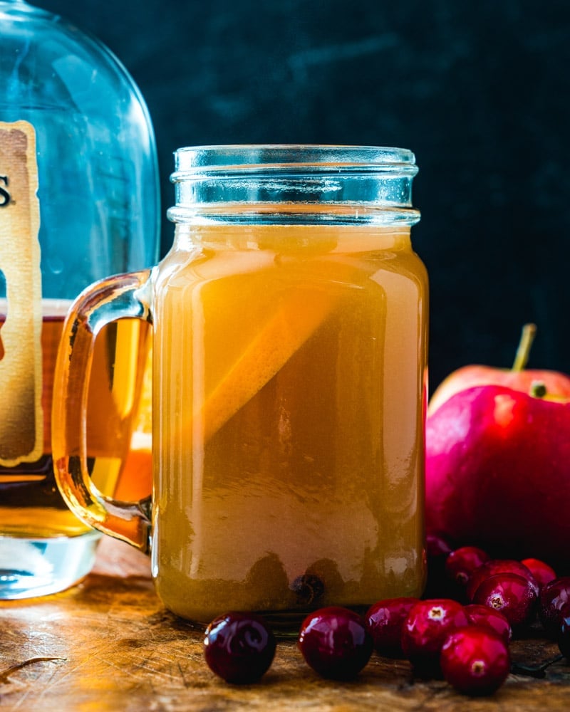 Spiked apple cider in a mason jar glass, surround by fruits, including apples and cherries, sitting on a wooden table