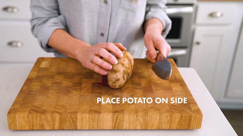 A potato is placed on its side on a cutting board, held by a woman's hand and a large knife in her opposite hand. It says, "Place potato on side."