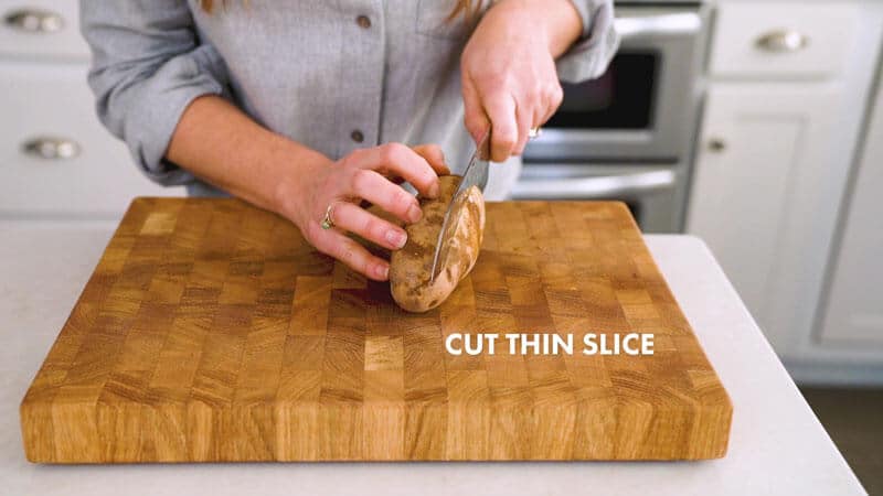 A potato is placed on its side on a cutting board, and a woman is positioning her knife to cut a thin slice into it. It says, "Cut thin slice."