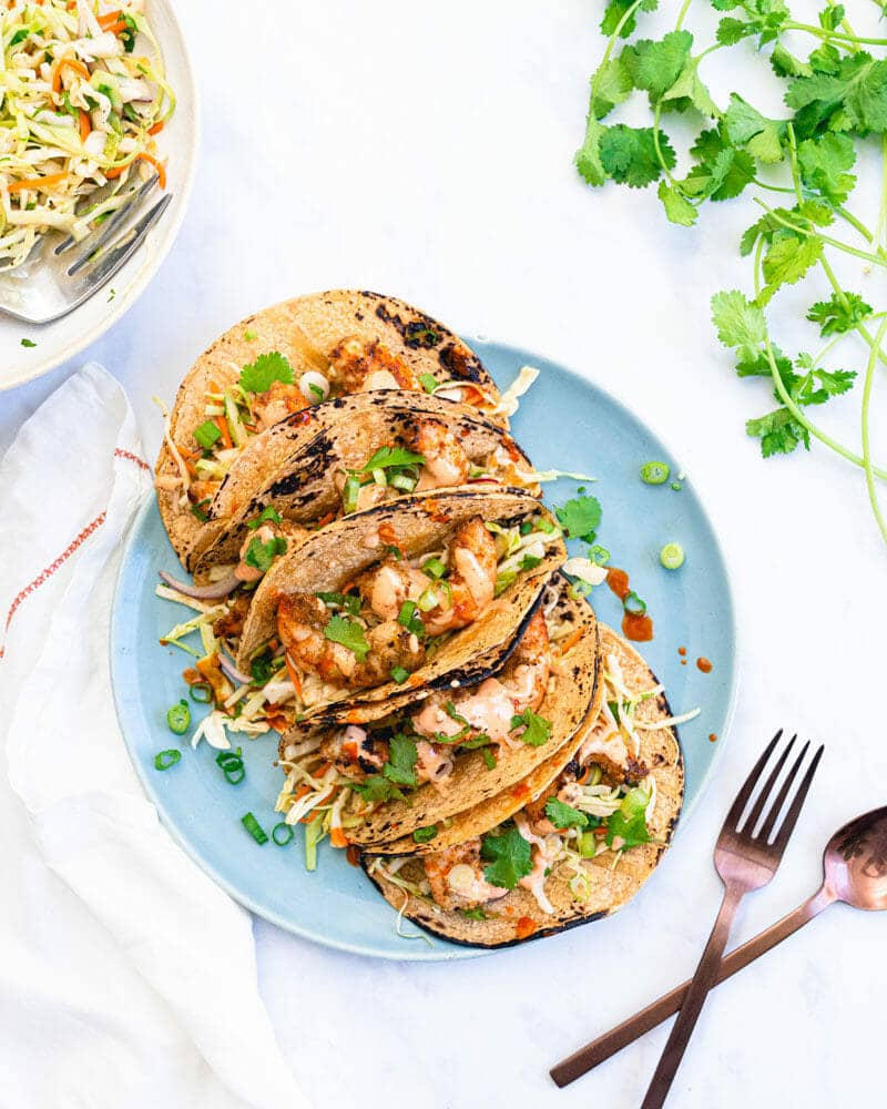 Several grilled shrimp tacos lined up on a plate and garnished with cilantro, surround by cilantro, utensils, and a bowl of coleslaw