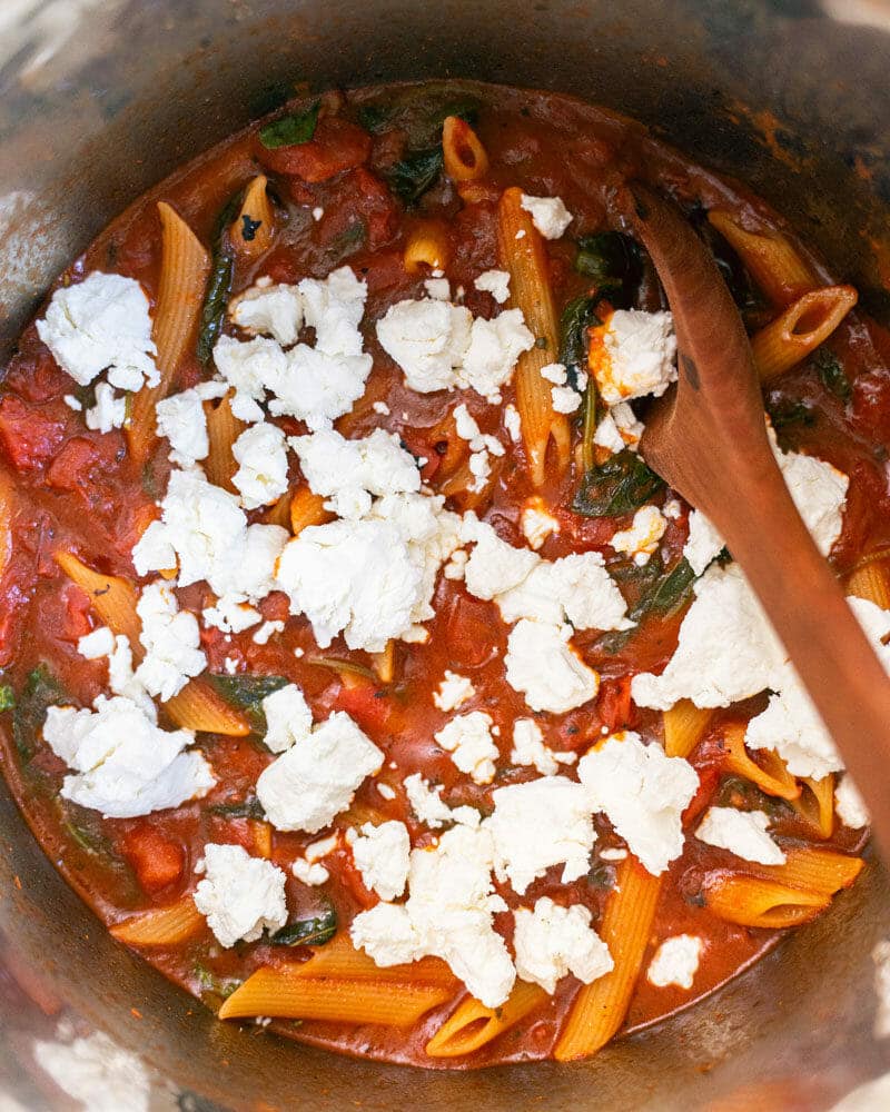 Pasta, tomatoes, goat cheese, and basil together in the Instant Pot and being mixed with a wooden spoon
