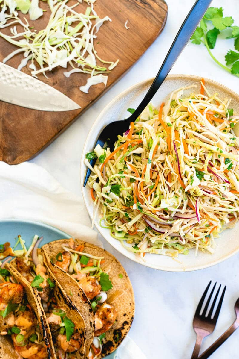 A colorful bowl of Mexican coleslaw with a fork on the plate, sitting near a cutting board with a knife and a plate with tacos