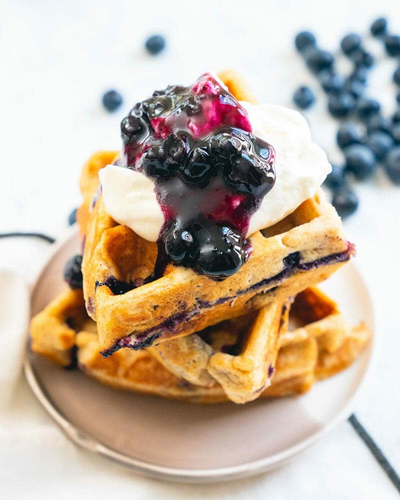 Blueberry waffles stacked on a plate and topped with whipped cream and blueberry sauce