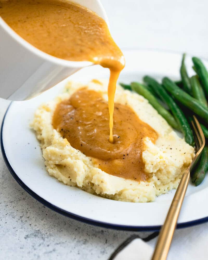Golden vegetarian gravy being poured over mashed potatoes on a plate next to some greens