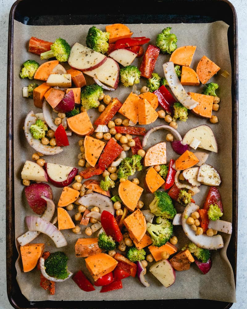 Sheet pan dinner lined with parchment paper and cover with red onions, sweet potatoes, brocoli, red peppers, and chickpeas