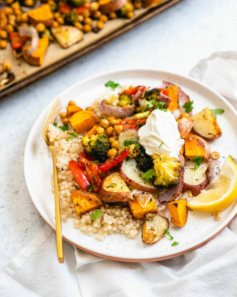 A sheet pan dinner on a plate served over couscous and topped with a mixture of veggies and chickpeas, and a lemon wedge for garnish