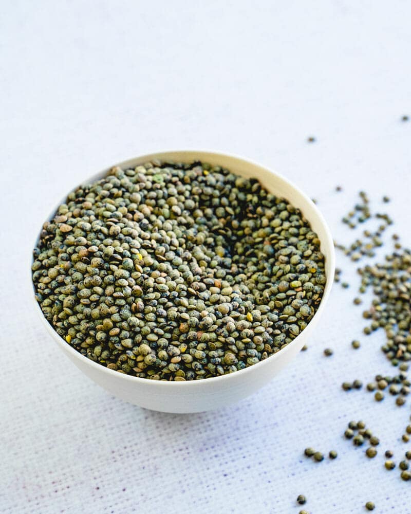 Uncooked French lentils in a bowl with more lentils on the counter surrounding