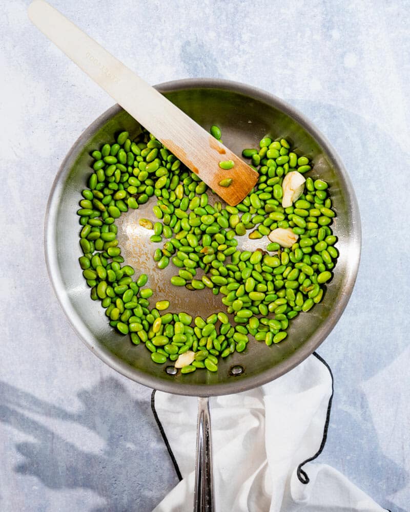 Edamame in pan with garlic and a wooden cooking utensil