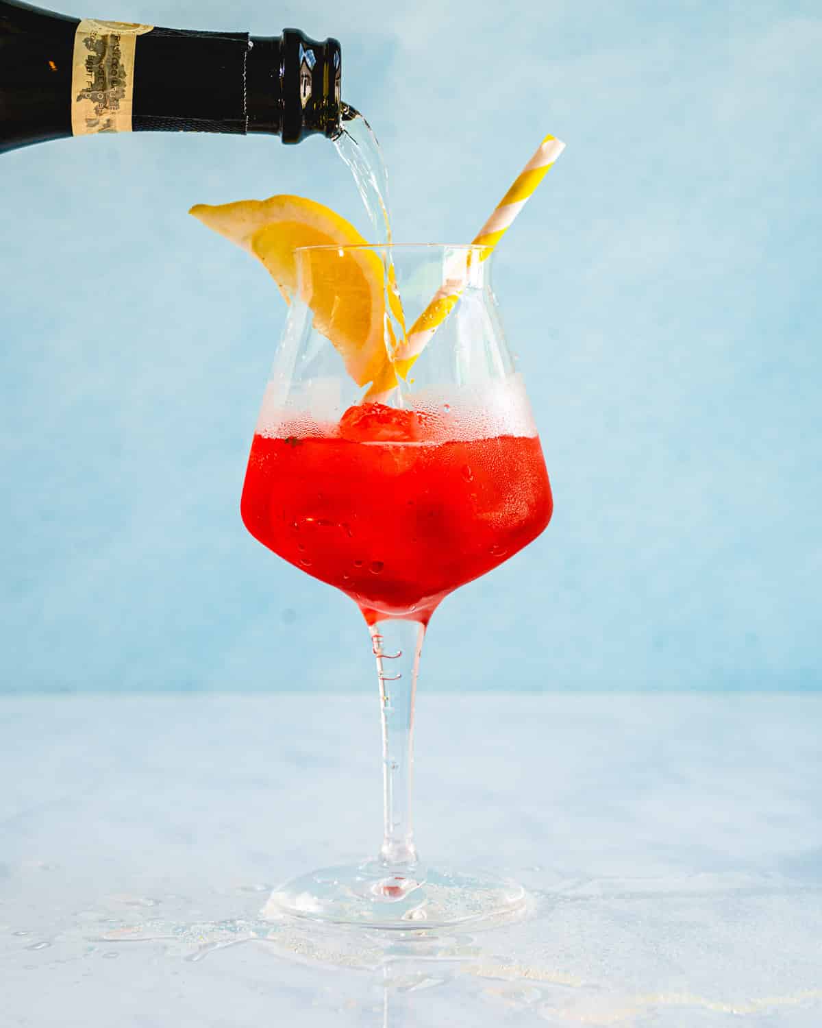 Campari spritz in a glass with a lemon wedge and a straw with  prosecco being poured in