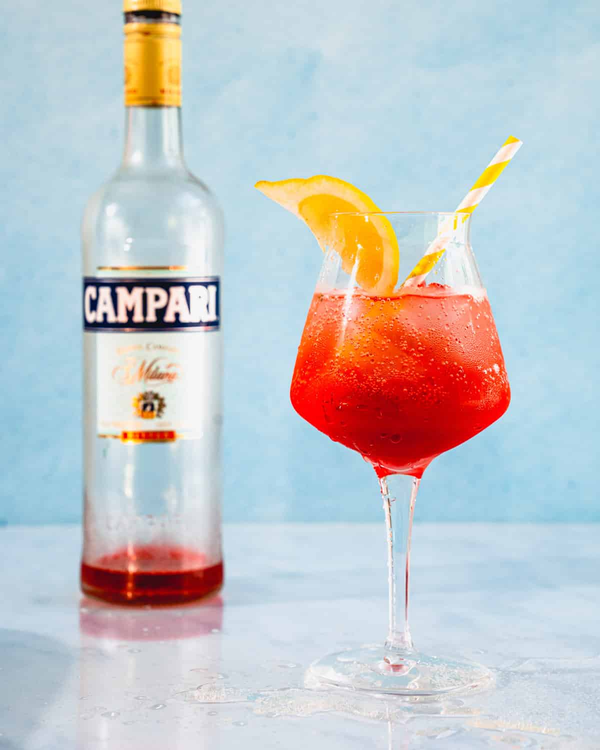 Campari spritz in a glass with a lemon wedge and a straw next to an almost empty bottle of Campari