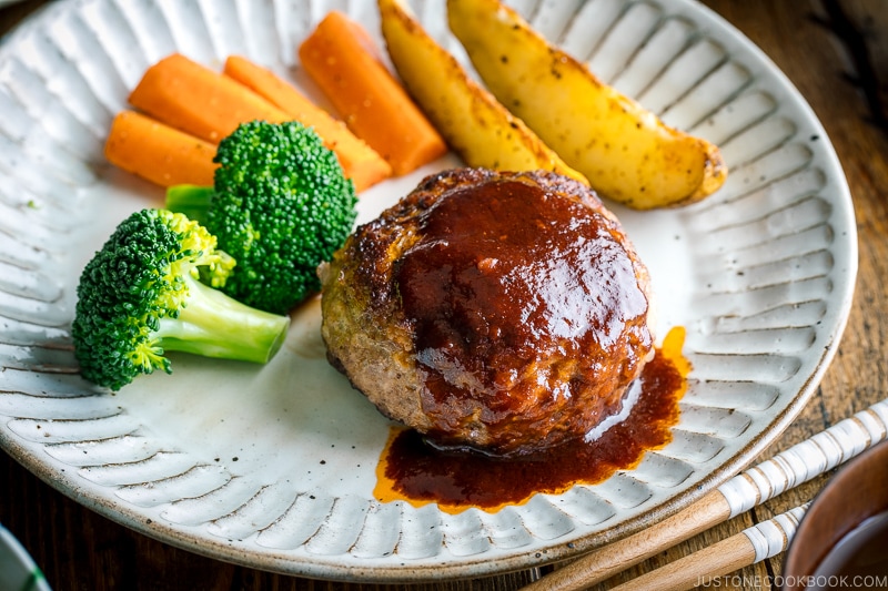 A white plate containing Japanese Hamburger Steak (Hambagu), saut&eacute;ed carrot, broccoli, and baked potato wedges.