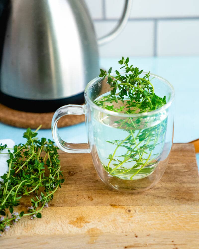 A clear mug with thyme and water sitting on a chopping board with fresh thyme next to it.