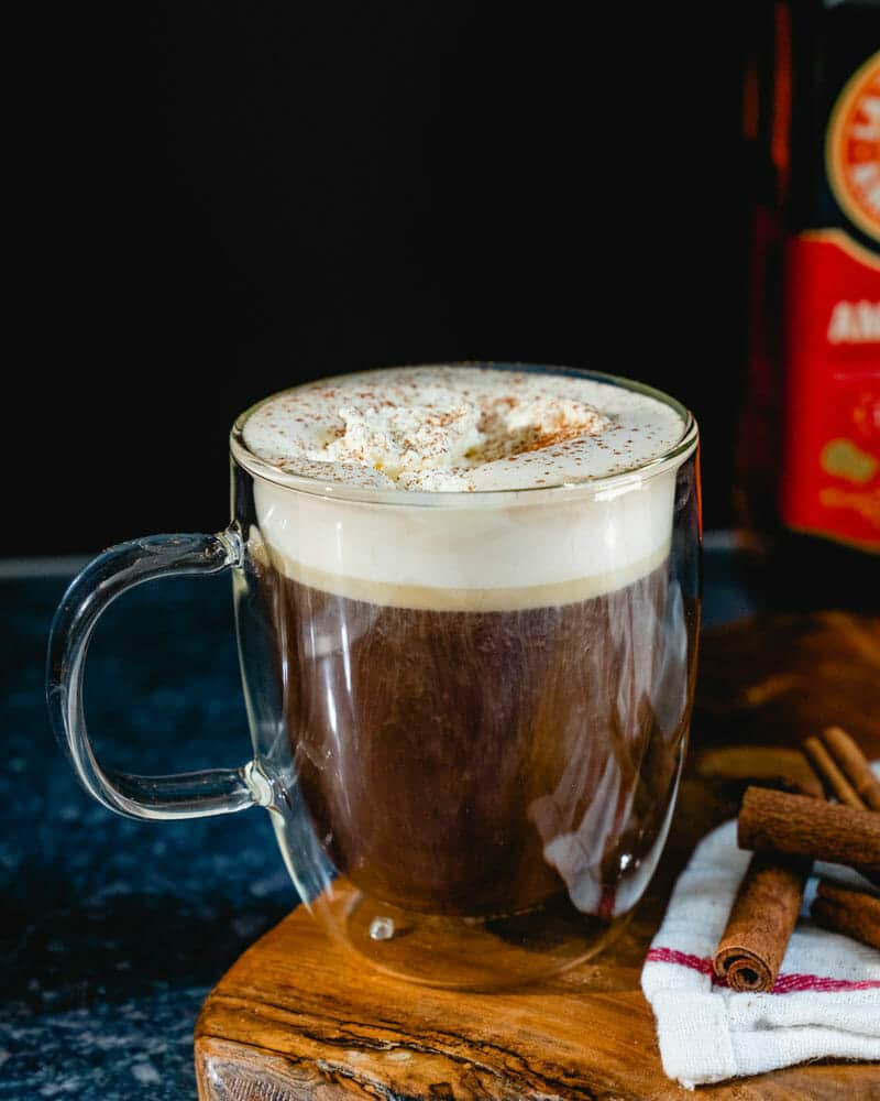 Amaretto coffee with whipped cream on top, sprinkled with cinnamon in a clear mug sitting next to stacked cinnamon sticks