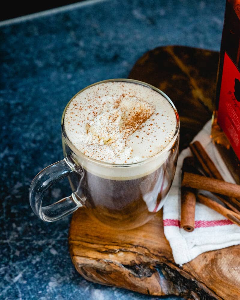 Amaretto coffee with whipped cream on top, sprinkled with cinnamon in a clear mug sitting next to stacked cinnamon sticks