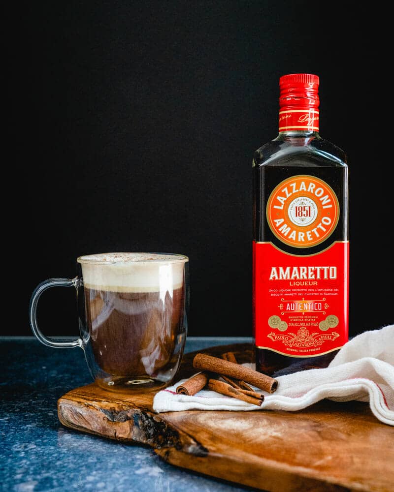 Amaretto coffee with whipped cream on top, sprinkled with cinnamon in a clear mug sitting next to stacked cinnamon sticks and a bottle of Amaretto Liqueur