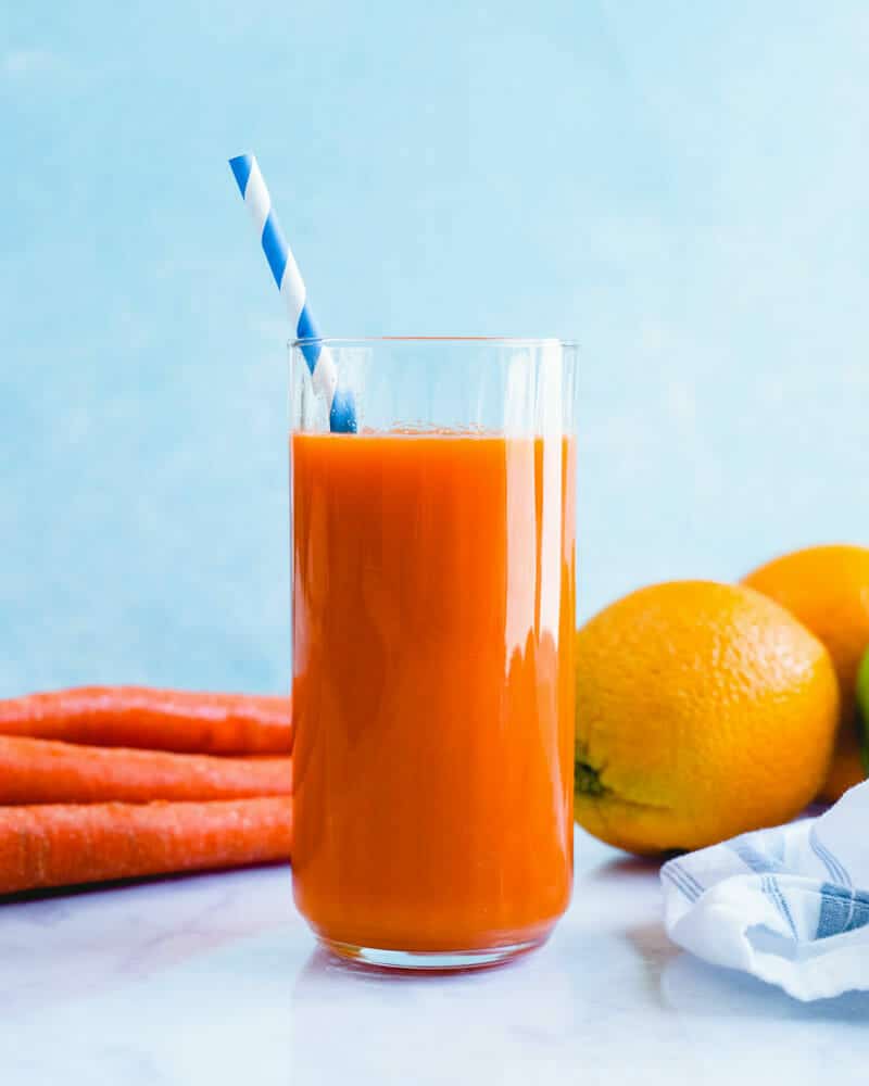 Carrot juice in a tall cup with a colorful straw, and carrots and oranges in the background