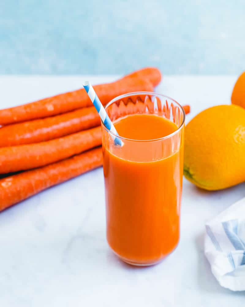 Carrot juice in a tall cup with a colorful straw, and carrots and oranges in the background