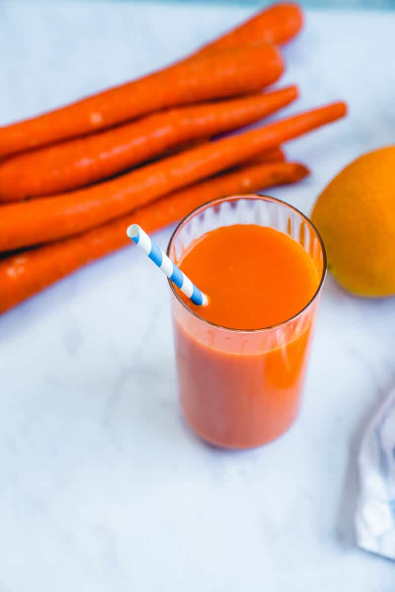 Top view of carrot juice in a tall cup with a colorful straw, and carrots and oranges in the background