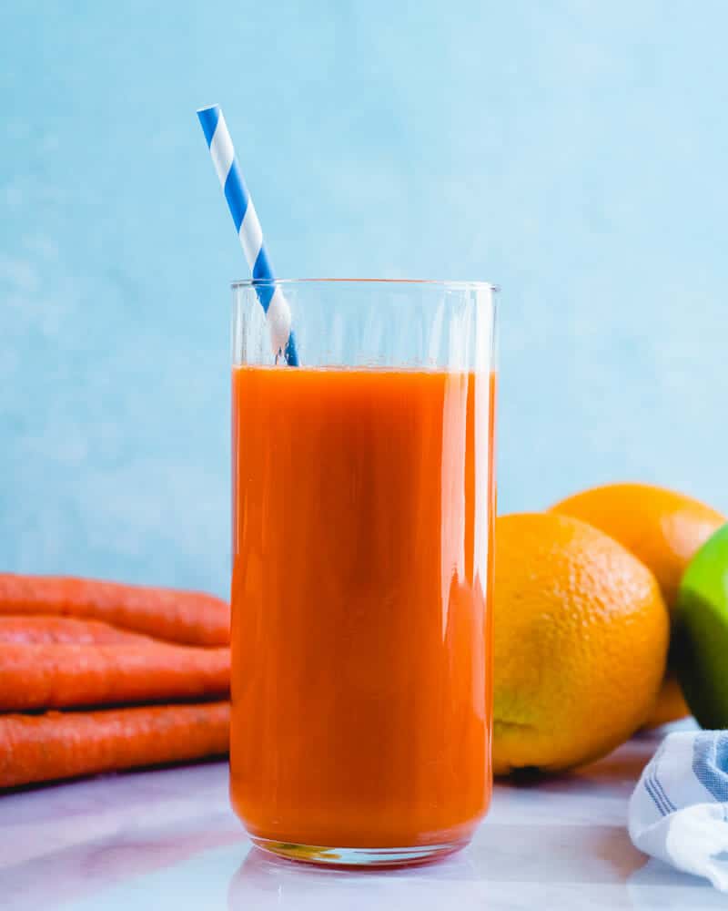 Carrot juice in a tall cup with a colorful straw, and carrots and oranges in the background