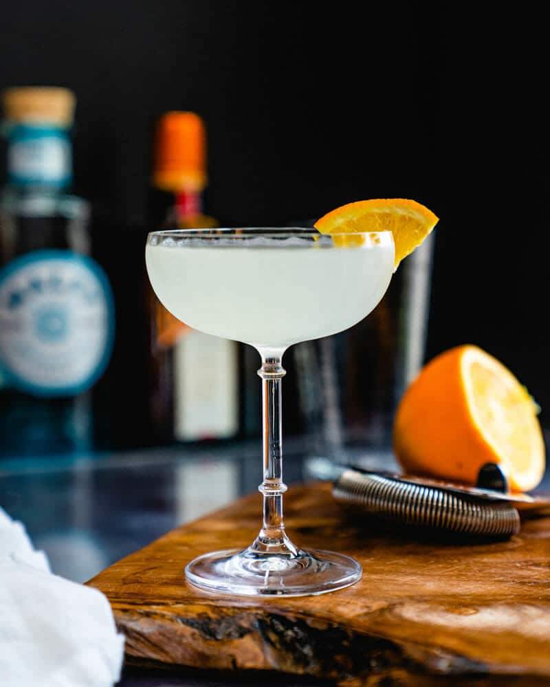 Corpse reviver No 2 cocktail in a tall glass and garnished with a lemon wedge and a half of a lemon in the background