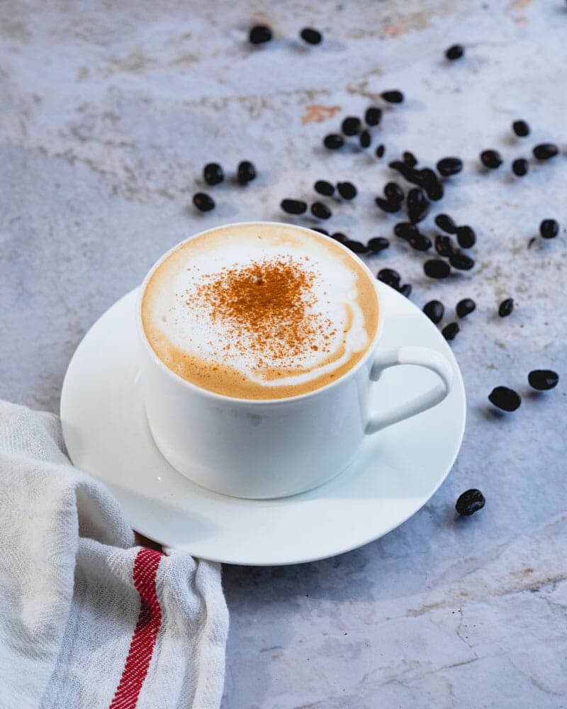 A cappuccino in a white cup and plate, surrounded by coffee beans, and sprinkled with cinnamon