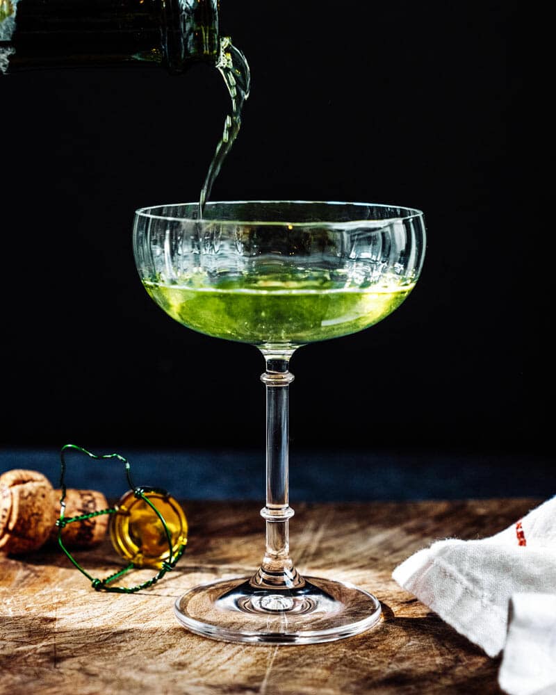 Death in the afternoon cocktail in a tall glass with a dark background and a liquor being poured into the glass