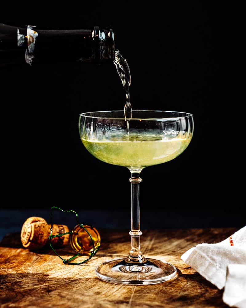 Death in the afternoon cocktail in a tall glass with a dark background and a liquor being poured into the glass