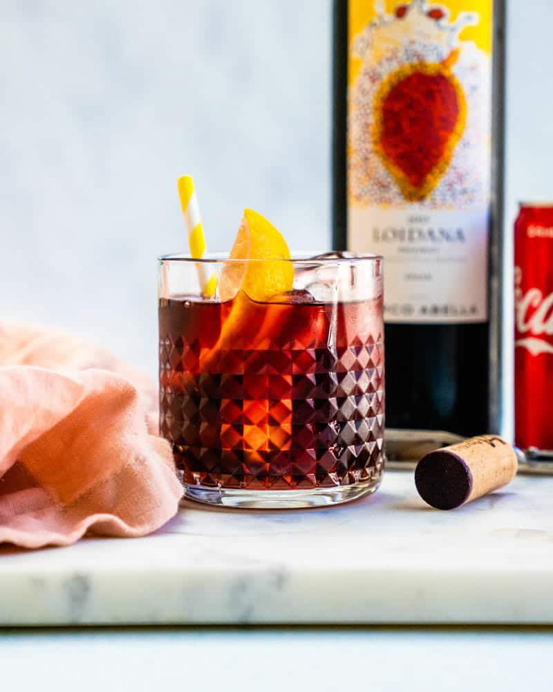 A short glass containing Kalimotxo over ice with a lemon wedge and colorful straw surrounded by a bottle of Loidana wine and Coca-Cola