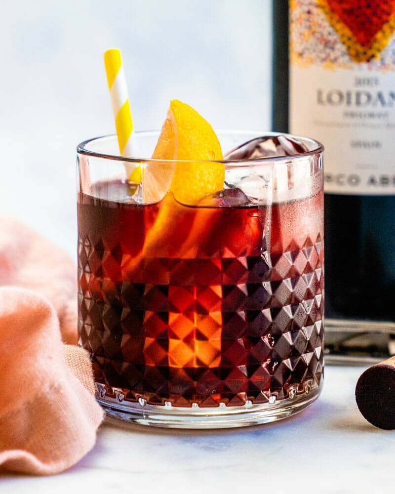 A short glass containing Kalimotxo over ice with a lemon wedge and colorful straw sitting on a table with a bottle of Loidana wine