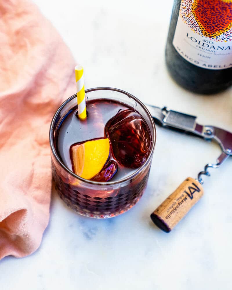 A short glass containing Kalimotxo over ice with a lemon wedge and colorful straw sitting on a table with a wine opener and bottle of Loidana wine