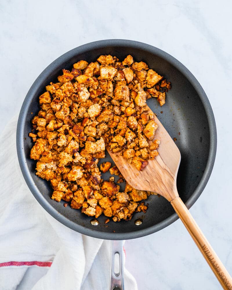 Tofu crumbles in a frying pan being tossed by a wooden cooking utensil