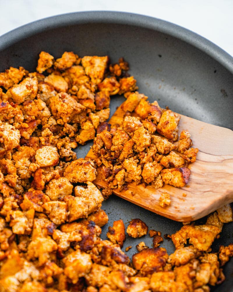 Tofu crumbles in a frying pan with a wooden cooking utensil