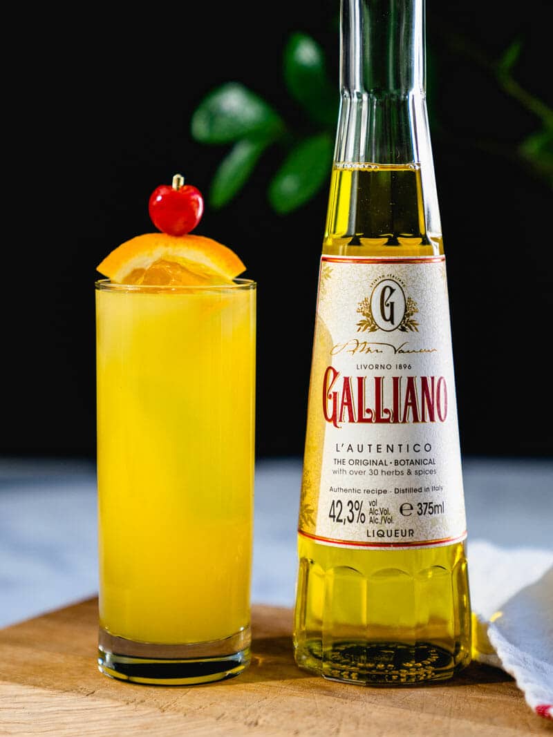 Harvey Wallbanger in a tall glass, garnished with an orange and cherry, sitting next to a bottle of Galliano
