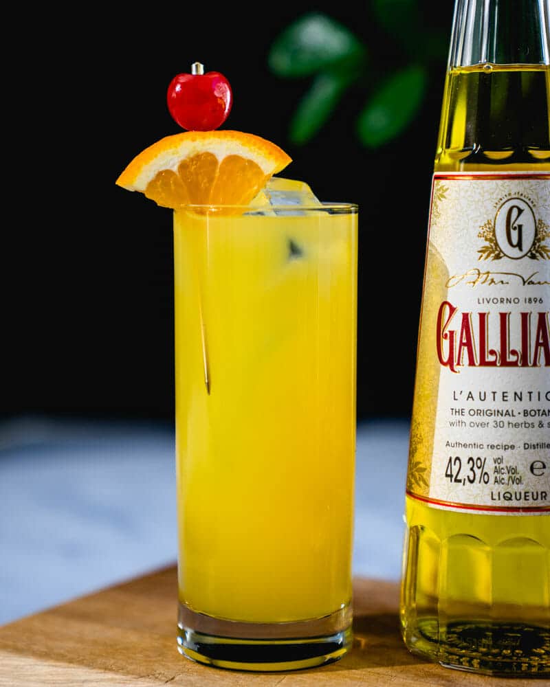 Harvey Wallbanger in a tall glass, garnished with an orange and cherry, sitting next to a bottle of Galliano