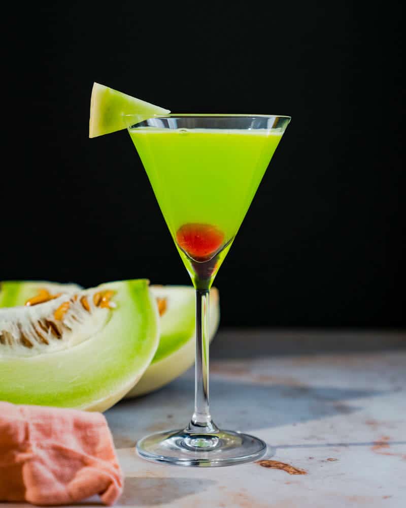 Japanese Slipper in a tall martini glass, garnished with a cherry and melon, and cut melons in the background