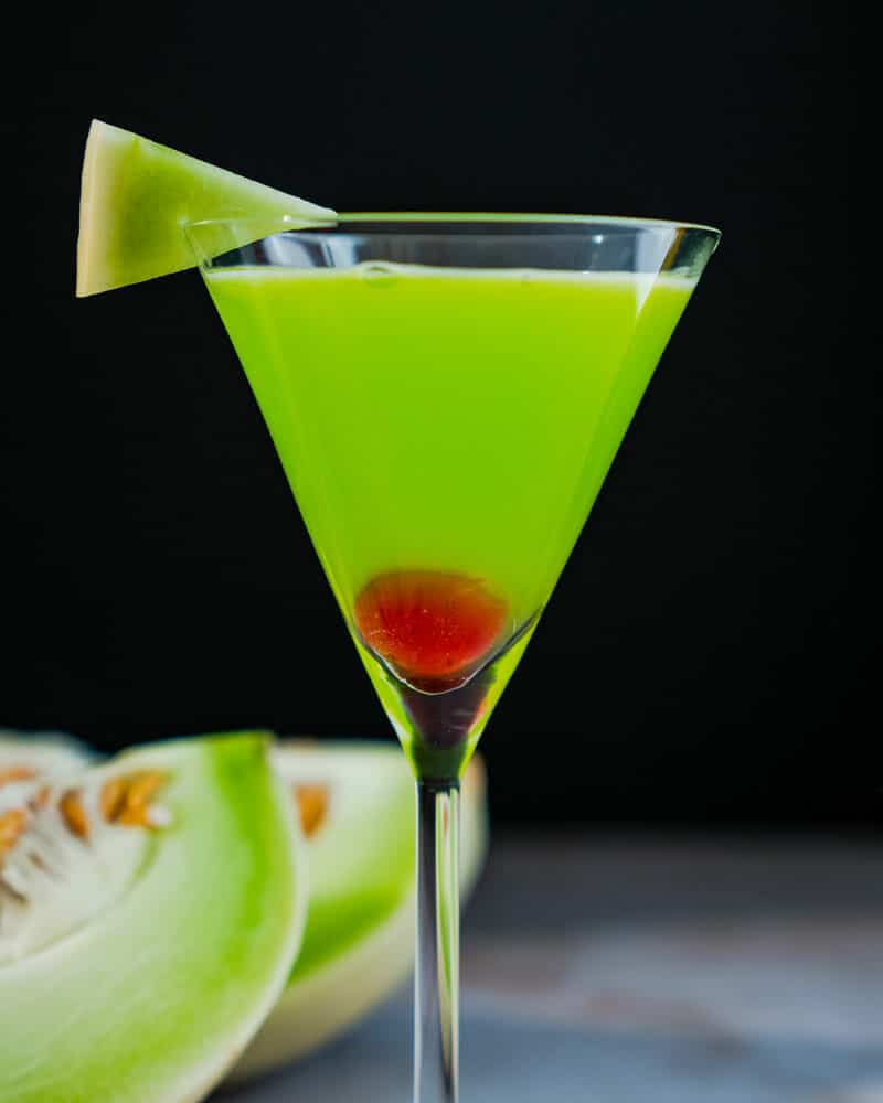 Japanese Slipper in a tall martini glass, garnished with a cherry and melon, and cut melons in the background