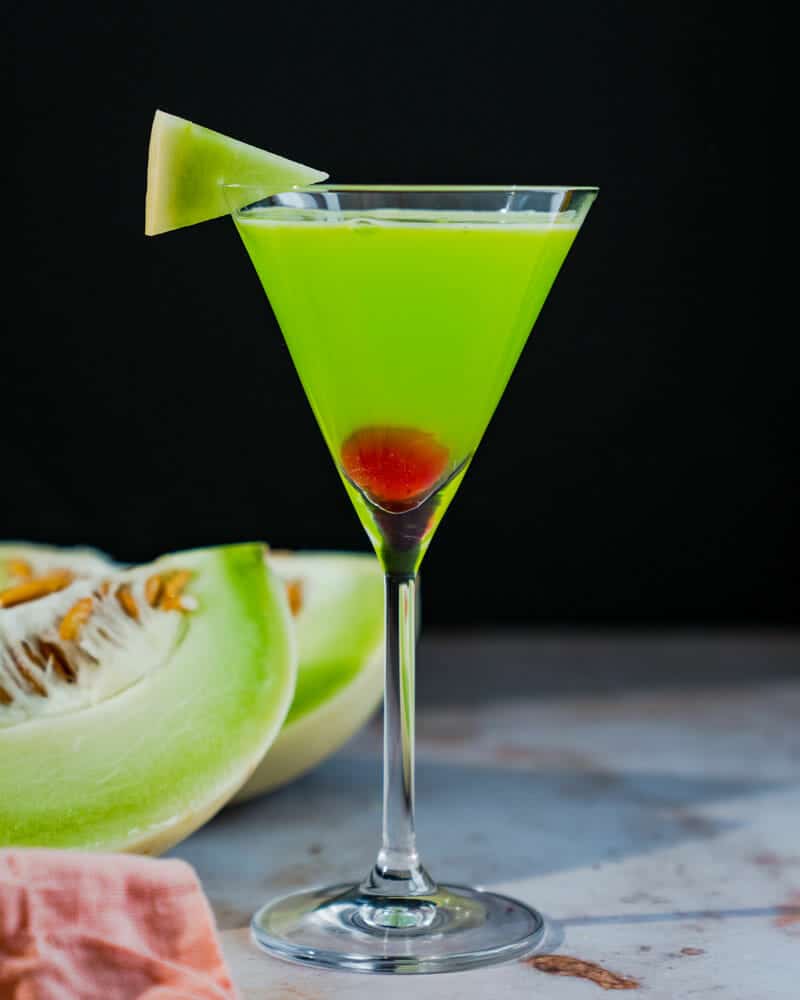 Japanese Slipper in a tall martini glass, garnished with a cherry and melon, and cut melons in the background