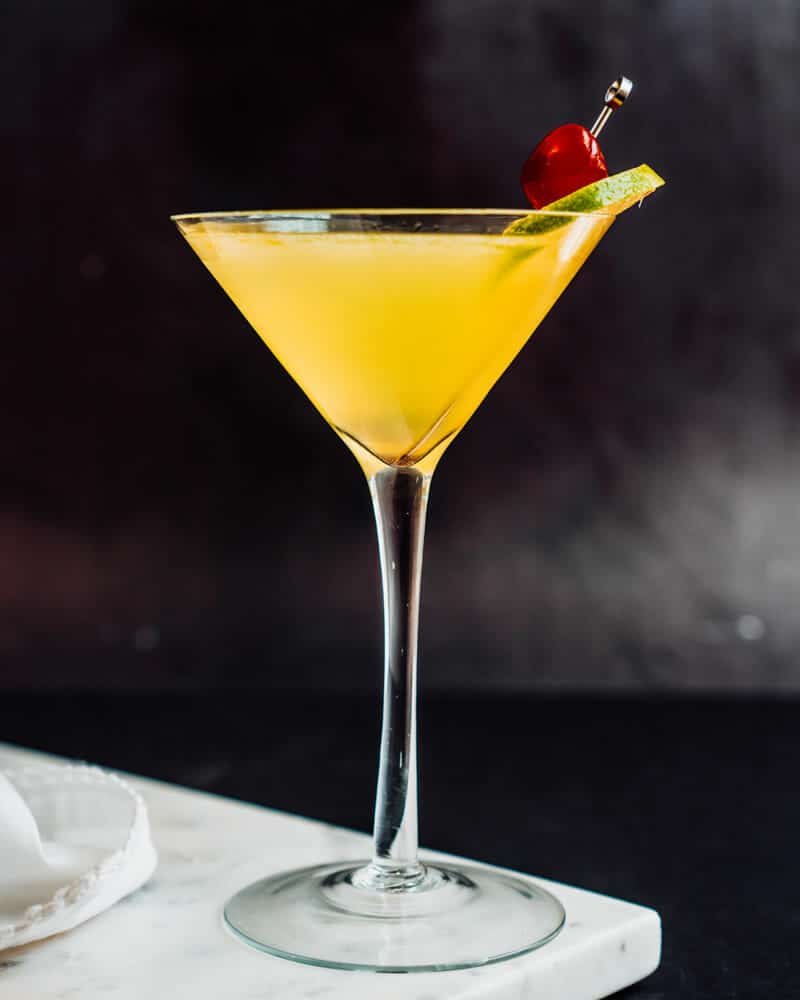 Yellow Bird Cocktail in a long-stemmed cocktail glass and garnished with lime and a cherry
