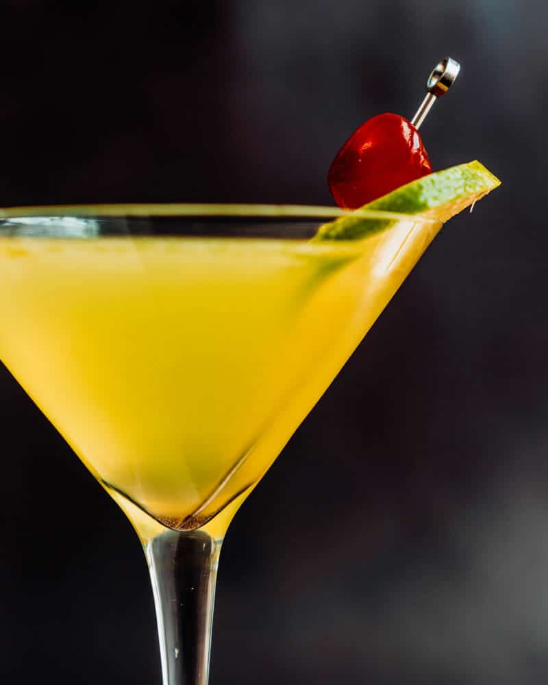 Yellow Bird drink up close in a martini glass. Garnished with lime and cherry
