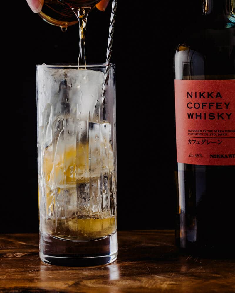 Whiskey being poured into a Japanese highball in a tall glass, garnished with a lemon, and sitting next to a bottle of Nikka Coffey Grain Whisky