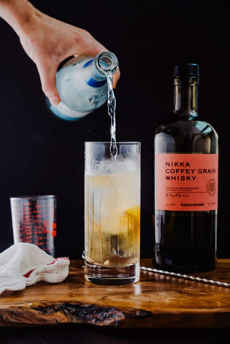 Club soda being poured into a Japanese highball in a tall glass, garnished with a lemon, and sitting next to a bottle of Nikka Coffey Grain Whisky