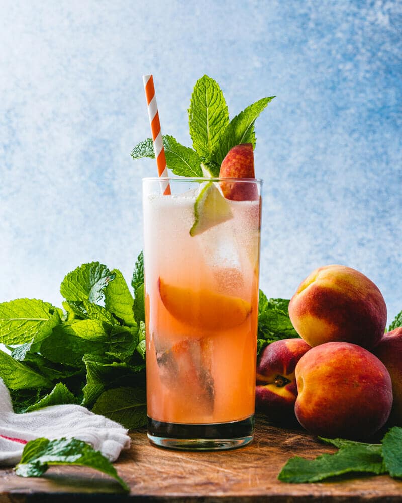 Peach cocktail with a colorful straw and garnishing surrounded by peaches sitting on a table.