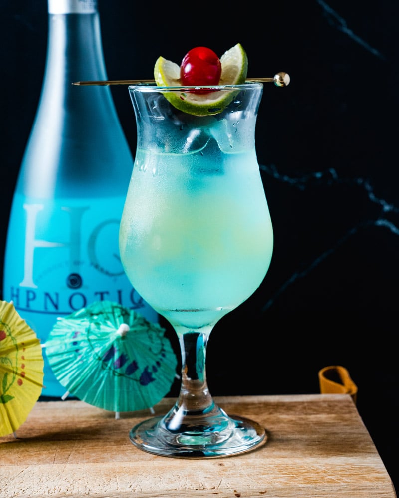 Hpnotiq Drink in a curvy glass with a garnished spear sitting across the top with a lime and cherry, surrounded by a bottle of Hpnotiq and small drink umbrellas