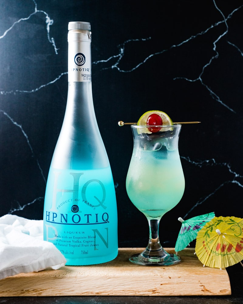 Hpnotiq Drink in a curvy glass with a garnished spear sitting across the top with a lime and cherry, surrounded by a bottle of Hpnotiq and small drink umbrellas