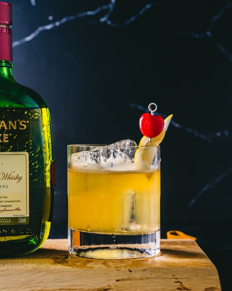 A Buchanan sour over ice in a short glass, garnished with lemon and cherry, sitting next to a bottle of Buchanan's