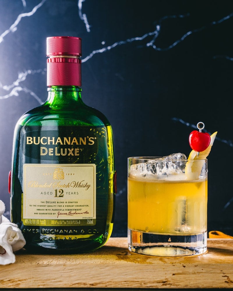 Buchanan sour in a lowball glass over ice and garnished with lemon and cherry, sitting next to a bottle of Buchanan's Deluxe